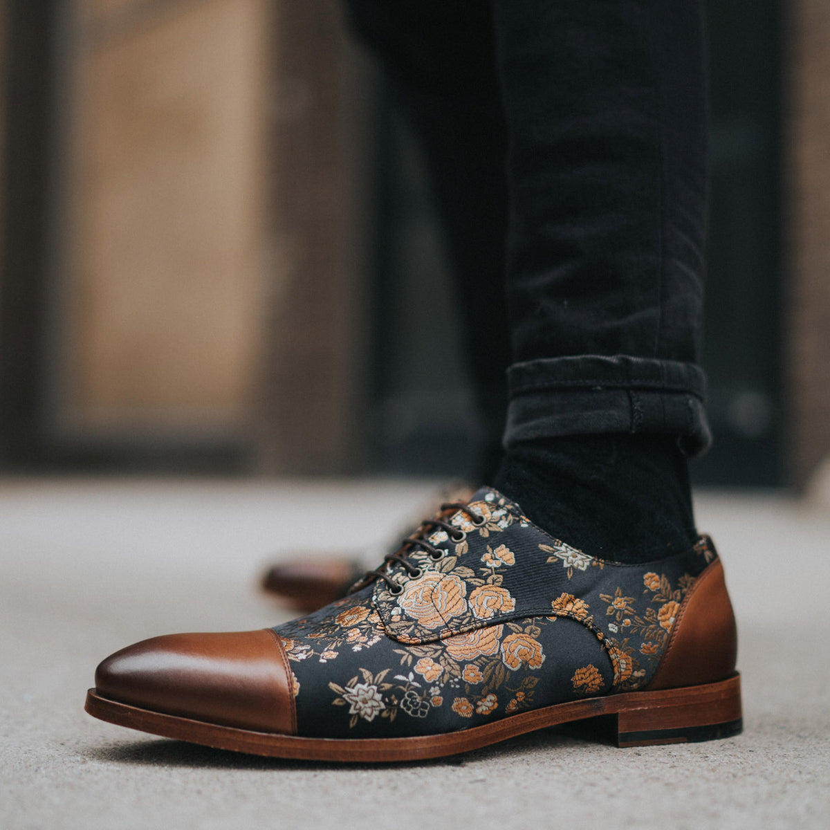 Close-up of a person wearing black pants and stylish shoes featuring a black base with gold floral patterns and brown leather toes and heels. The background is a blurred street scene.