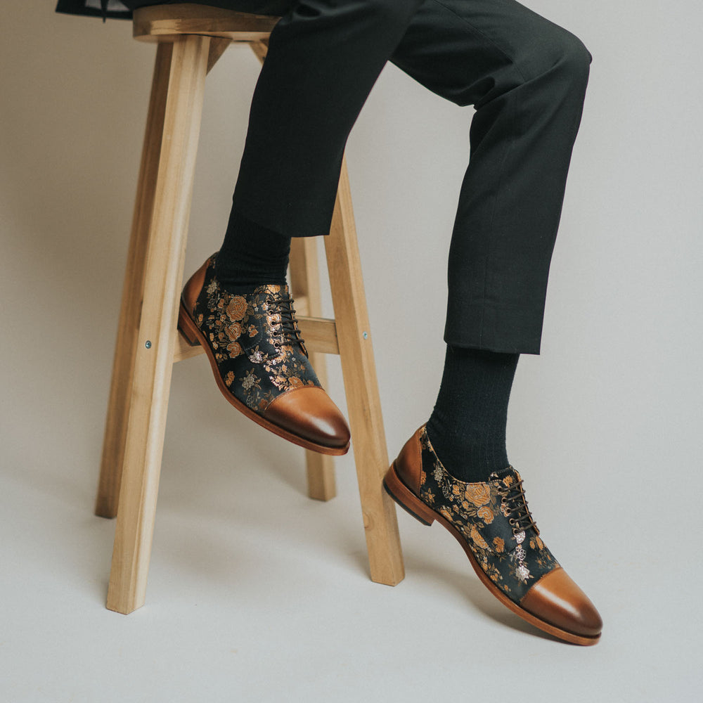 A person wearing black pants and stylish brown and black floral-patterned dress shoes sits on a wooden stool with crossed ankles.