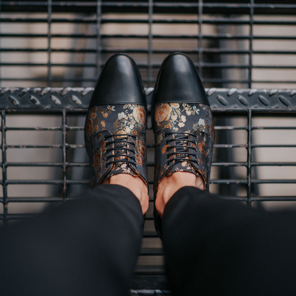 A pair of feet in black floral-patterned dress shoes stands on metal grating.