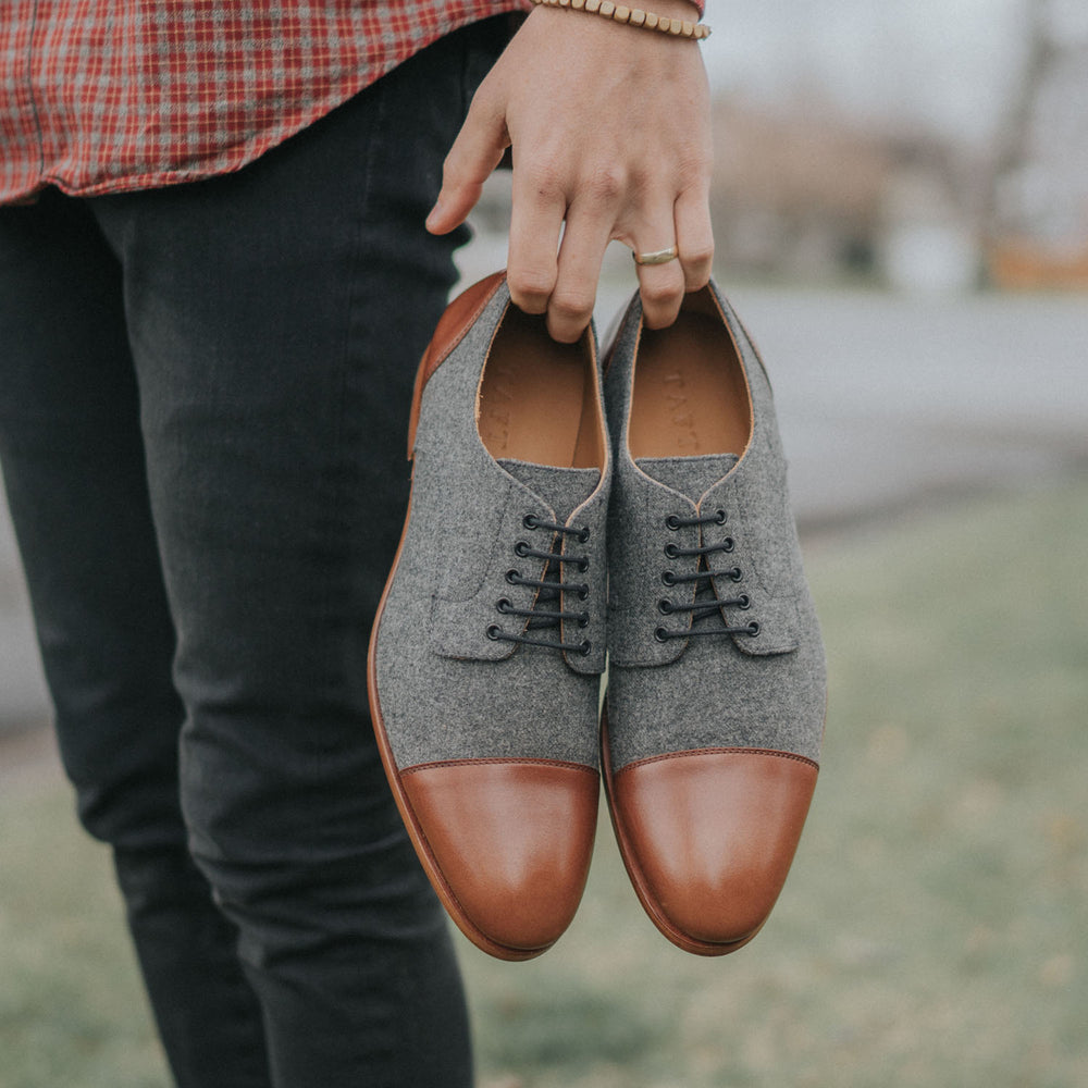 A person holds a pair of gray and brown dress shoes outdoors.