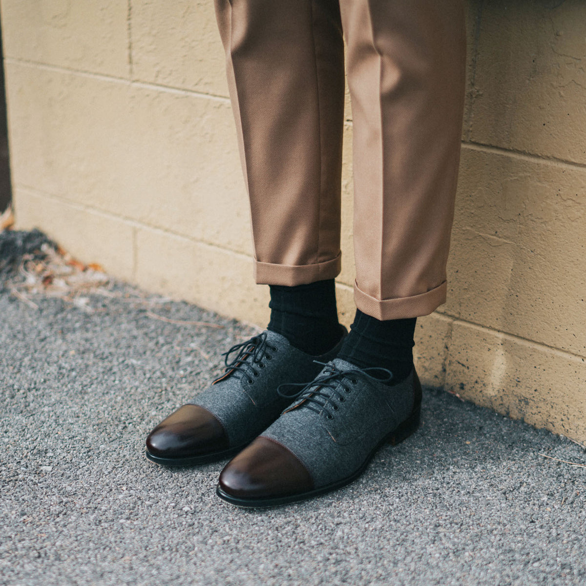 A person wearing brown pants, black socks, and gray dress shoes stands against a beige wall.