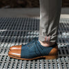 A person wearing gray pants with red stitching is seen from the knee down, displaying a pair of brown and blue oxford shoes on a metal grate surface.