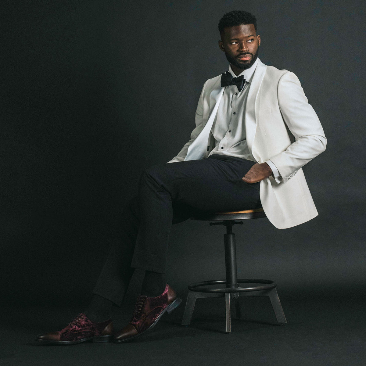 A man in a white blazer, black bow tie, and dark trousers sits on a stool against a dark background, looking slightly to the side.