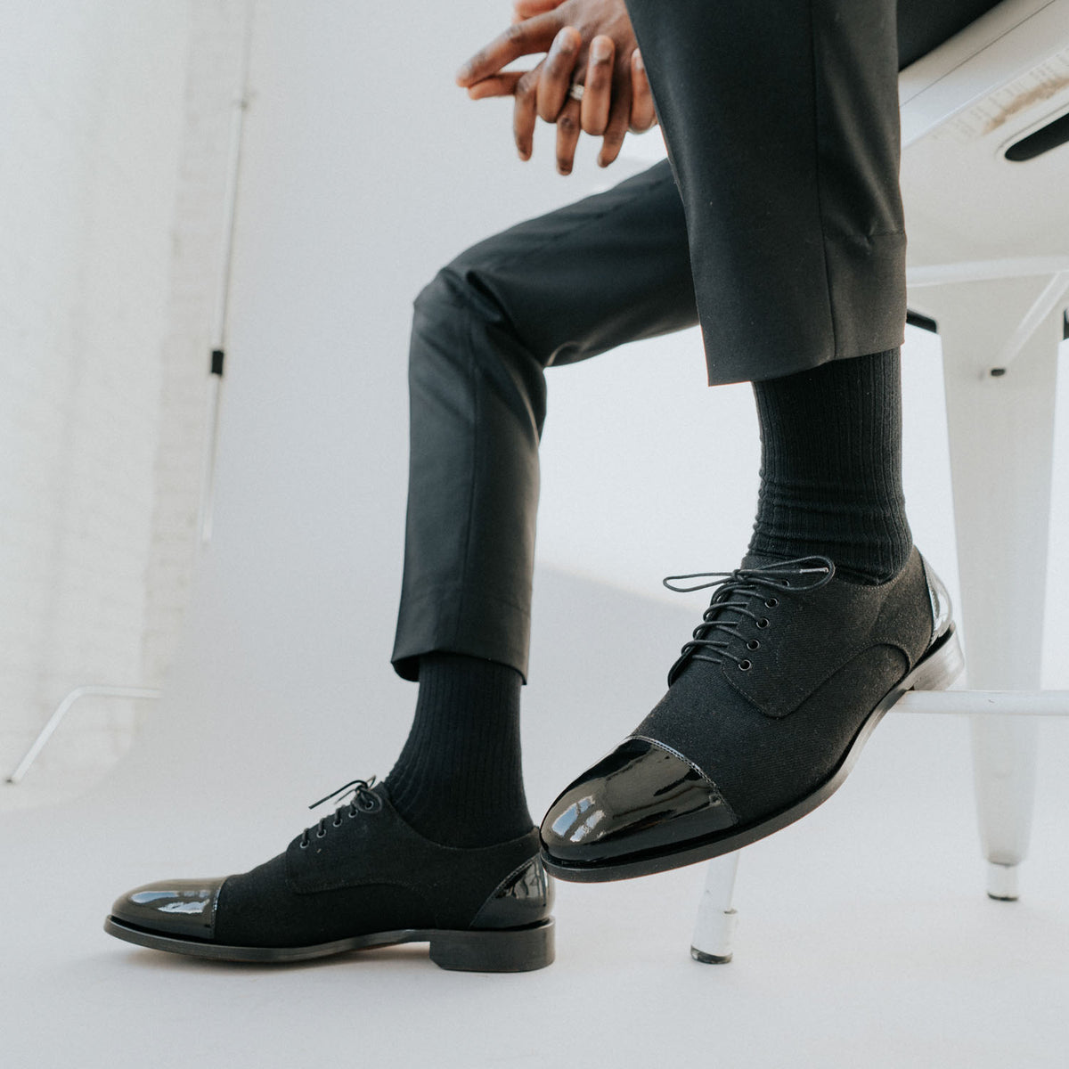 A person sits on a chair wearing black dress shoes with shiny cap toes, black socks, and black pants.