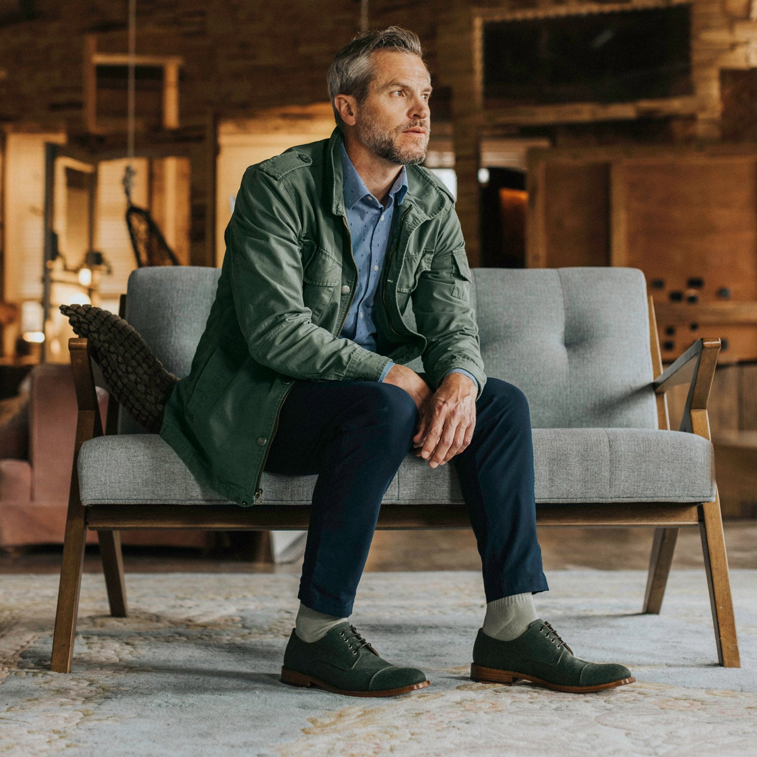 A man with gray hair sits on a gray sofa in a modern rustic interior, wearing a green jacket, blue shirt, blue pants, and green shoes, looking off into the distance.