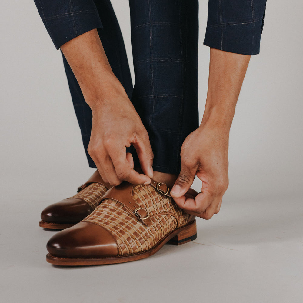 A person wearing navy pants adjusts the buckle on brown and beige woven dress shoes.