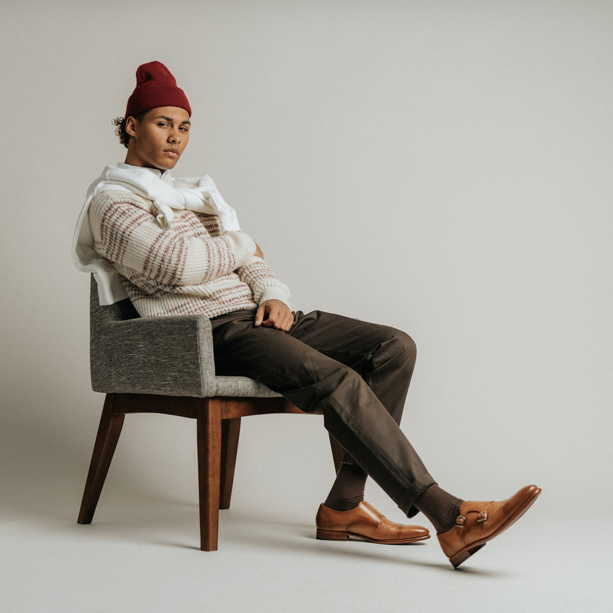 A person wearing a red beanie, white sweater, brown pants, and tan loafers sits on a gray chair with an arm draped over the backrest.