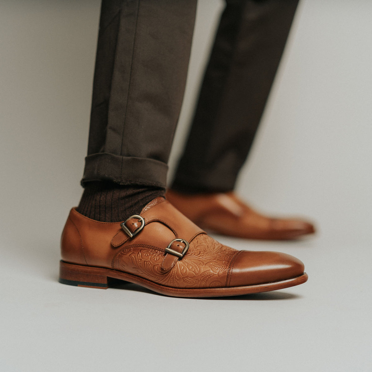 A person wearing brown leather double monk strap shoes with intricate detailing, dark brown socks, and dark pants.