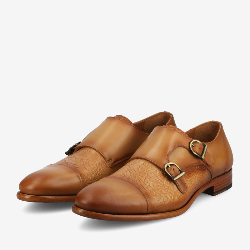 A pair of tan leather monk strap shoes with decorative engravings and two metal buckles on each shoe.