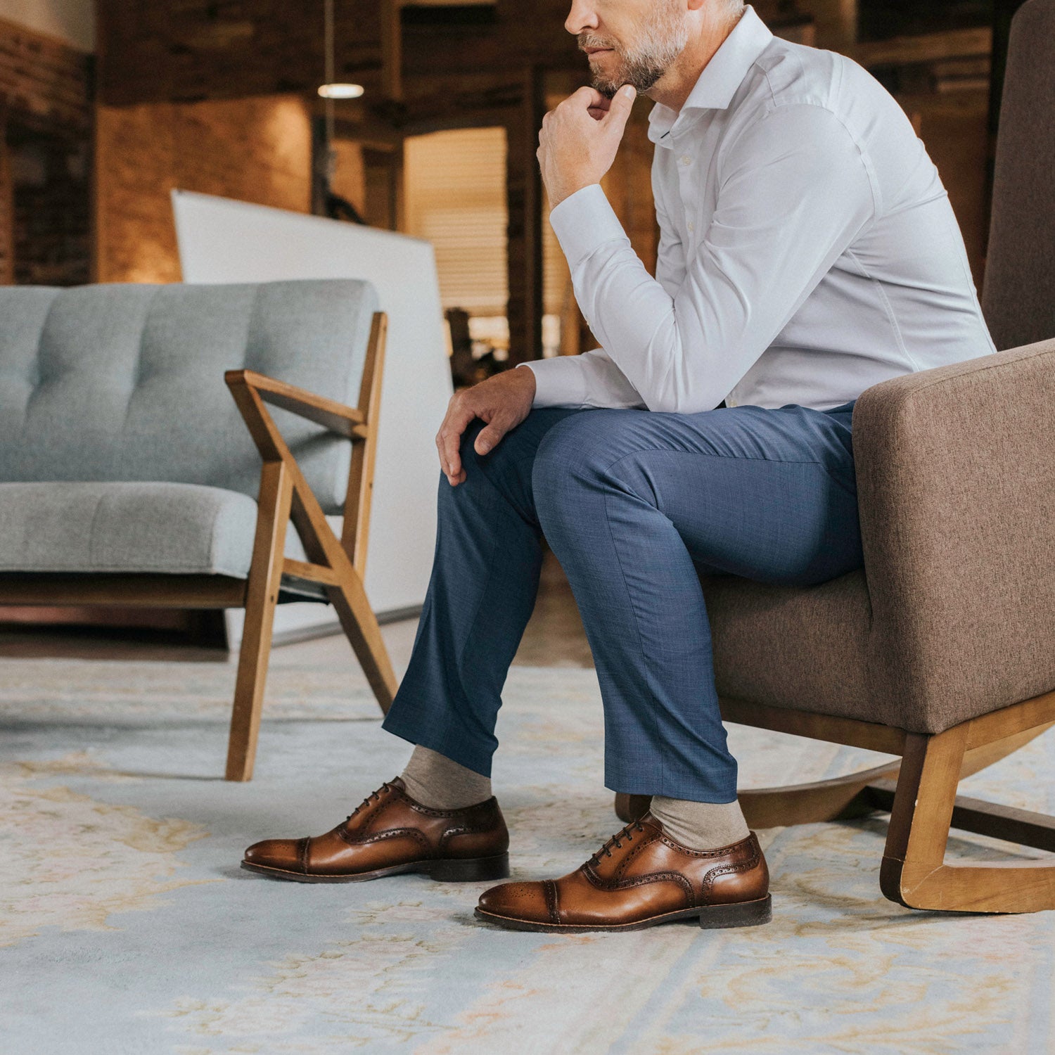 A man in a white shirt and blue trousers sits in an armchair, resting his chin on his hand, showcasing brown leather shoes.