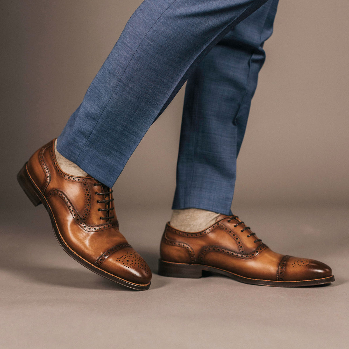 A person wearing brown leather dress shoes with perforated details and blue trousers.