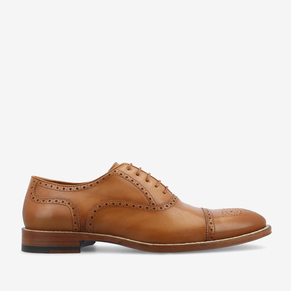 A tan leather oxford shoe with brogue detailing and a wooden sole viewed from the side.