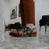 A person wearing rust-colored pants and floral-patterned shoes stands on a marble floor in a modern, art-adorned room.