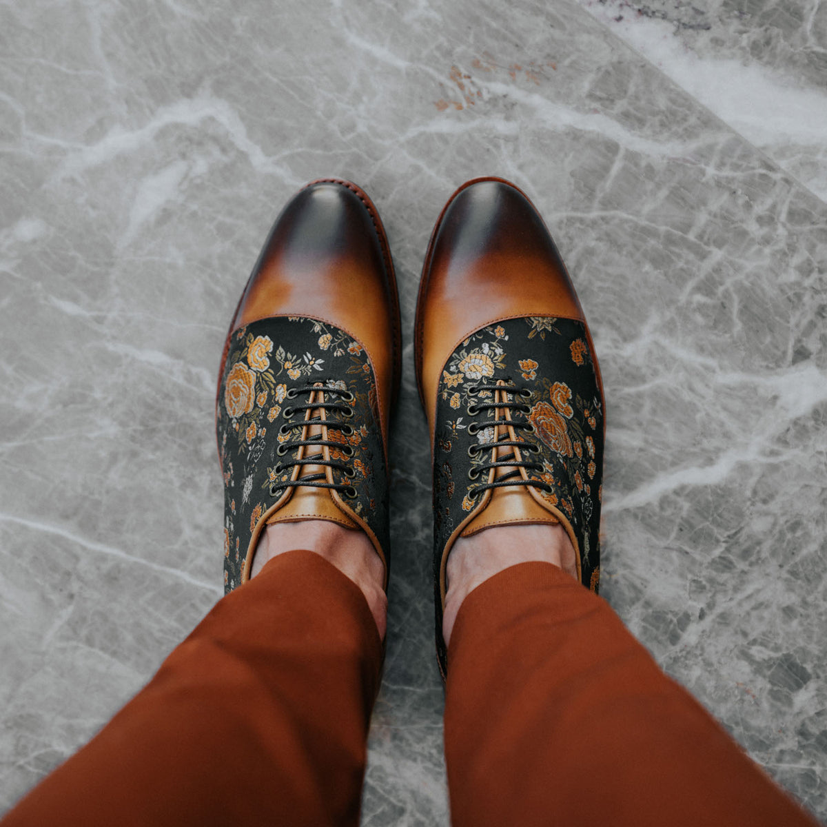 A person wearing brown and black oxford shoes with floral patterns, standing on a gray marble floor in brown pants.