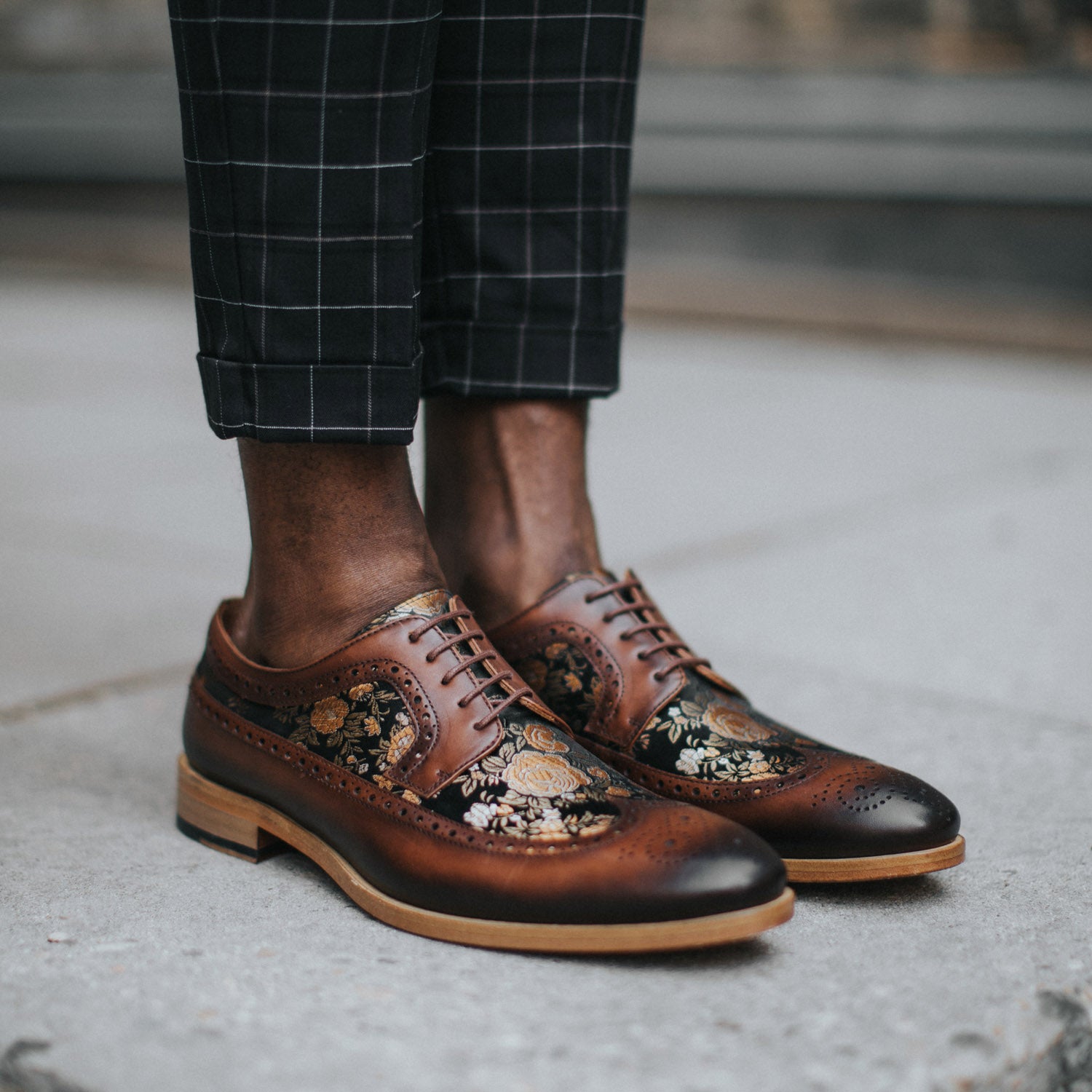 Close-up of a person wearing brown leather dress shoes with intricate floral designs, paired with black checkered pants.