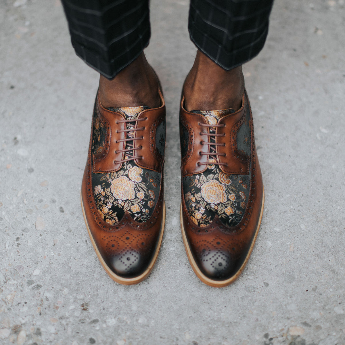 Close-up of a person wearing brown dress shoes with an intricate floral pattern, standing on a concrete surface.