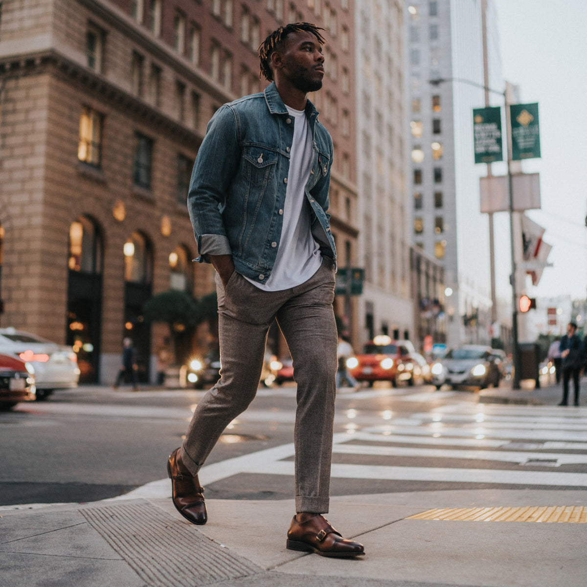 Man in a denim jacket and dress pants crossing a city street with tall buildings and cars in the background.