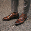 A person wearing brown leather monk strap shoes with plaid pants stands on a textured concrete surface.