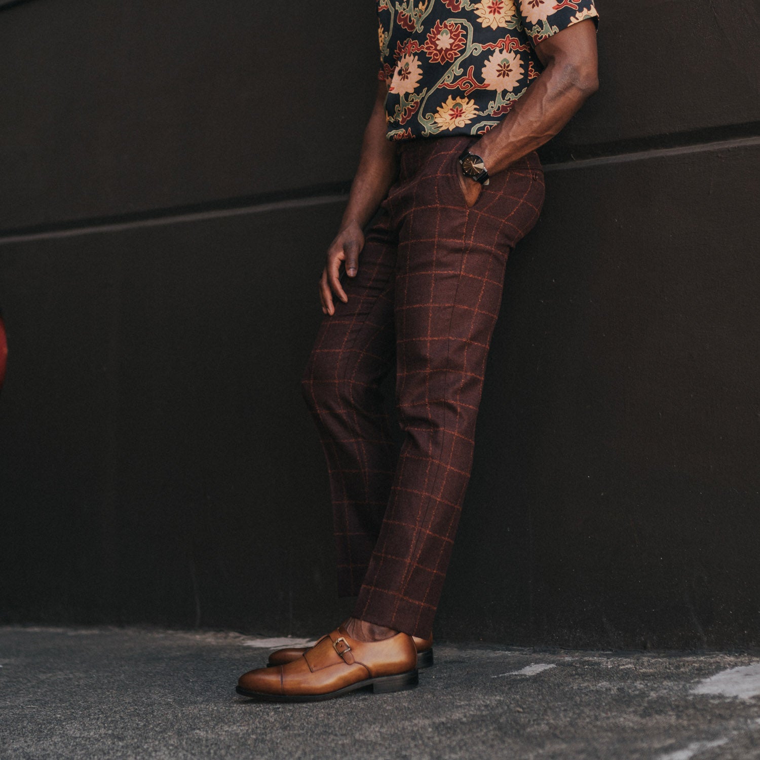 A person wearing a floral shirt, plaid pants, and tan dress shoes leans against a dark wall with hands in pockets.