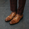 A person wearing brown checkered pants and tan double monk strap dress shoes stands on a concrete surface.