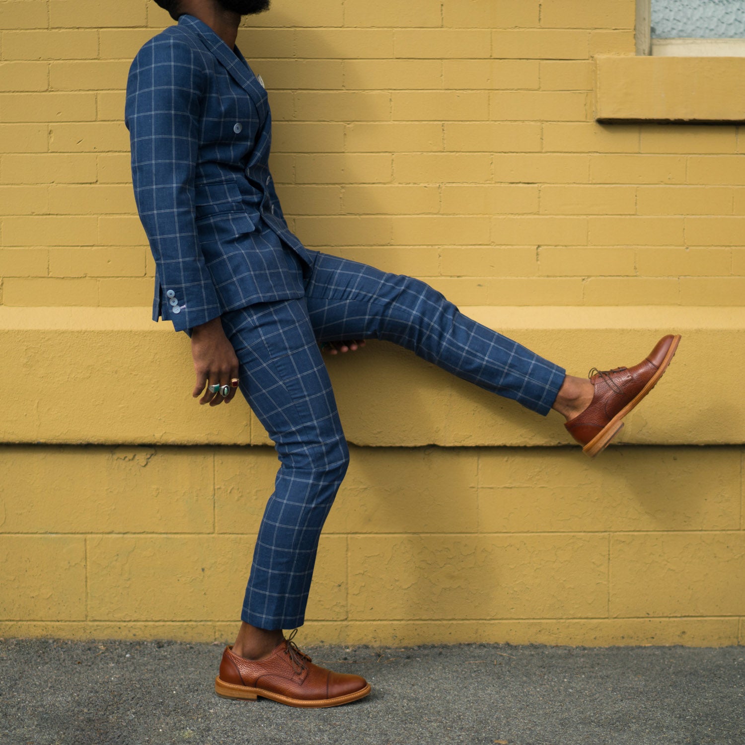 Person wearing a blue plaid suit with brown dress shoes walks against a yellow brick wall background.