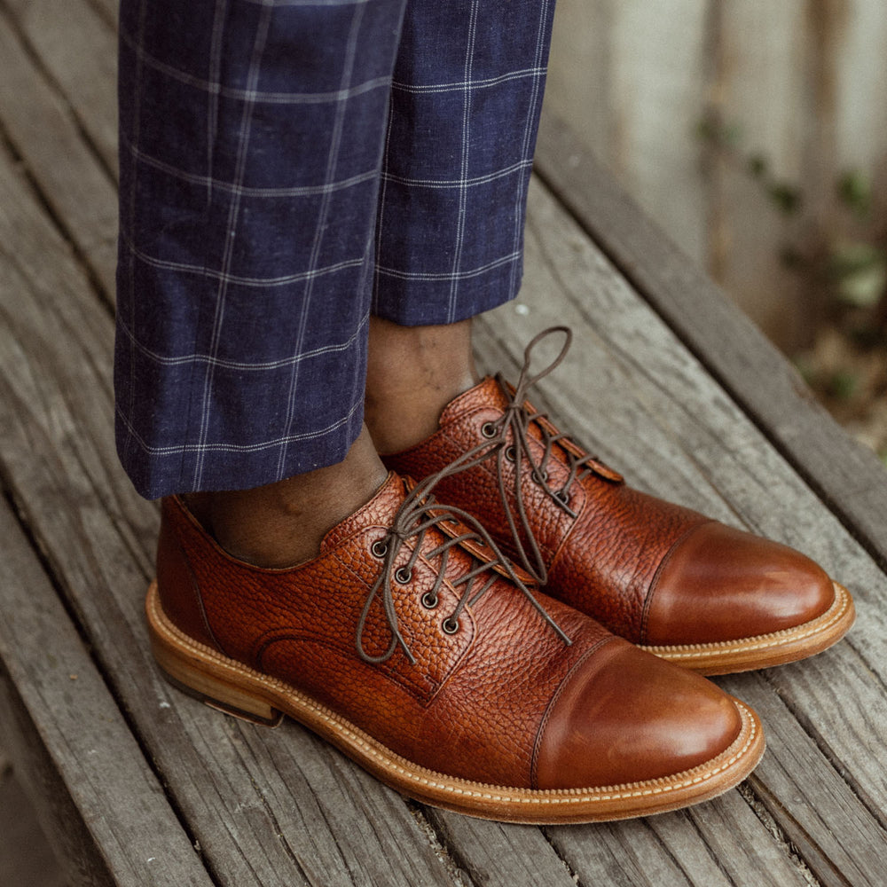 A person wearing brown leather shoes and blue checkered trousers stands on a wooden surface.