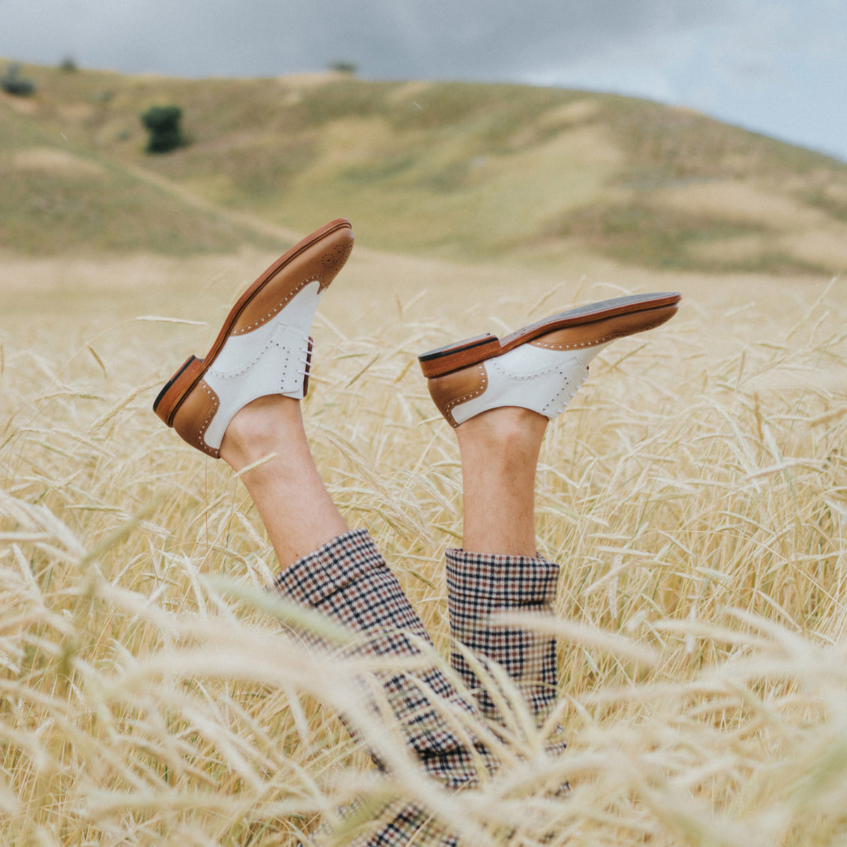 Legs in plaid pants sticking up from a field of tall dry grass, wearing white and brown shoes against a backdrop of rolling hills under a cloudy sky.