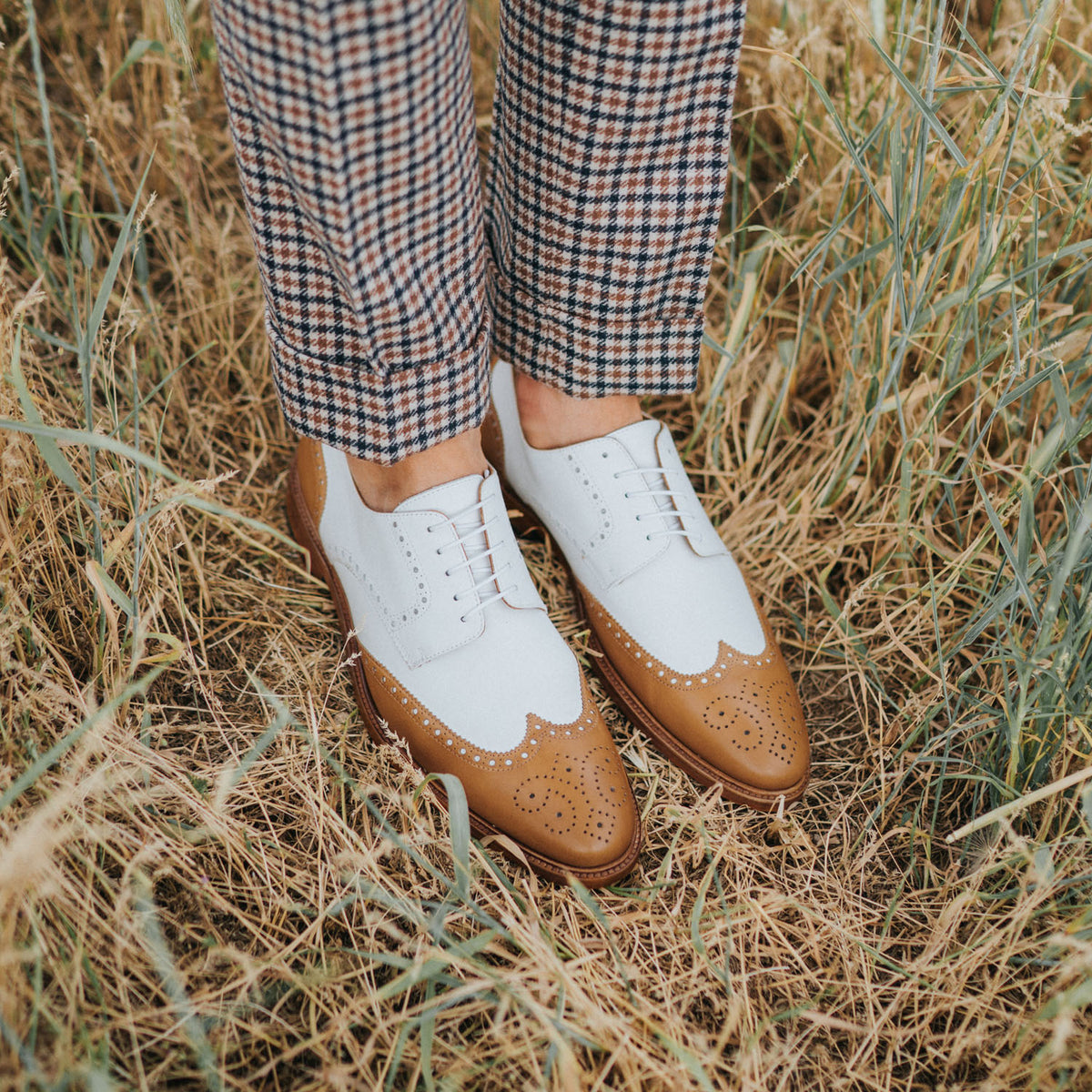 A person wearing checkered pants and white and brown wingtip shoes stands in tall dry grass.