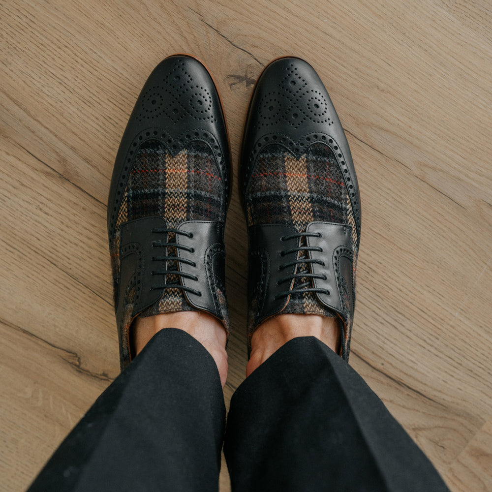 A pair of black and plaid leather dress shoes worn by a person, standing on a wooden floor.