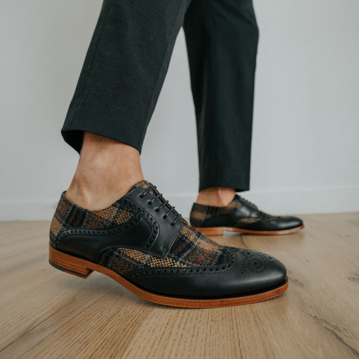 Close-up of a person wearing black and brown plaid oxford shoes with black trousers, standing on a wooden floor.