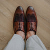 Brown leather and tweed oxford shoes with brogue detailing on a wooden floor, worn by a person in light gray pants.