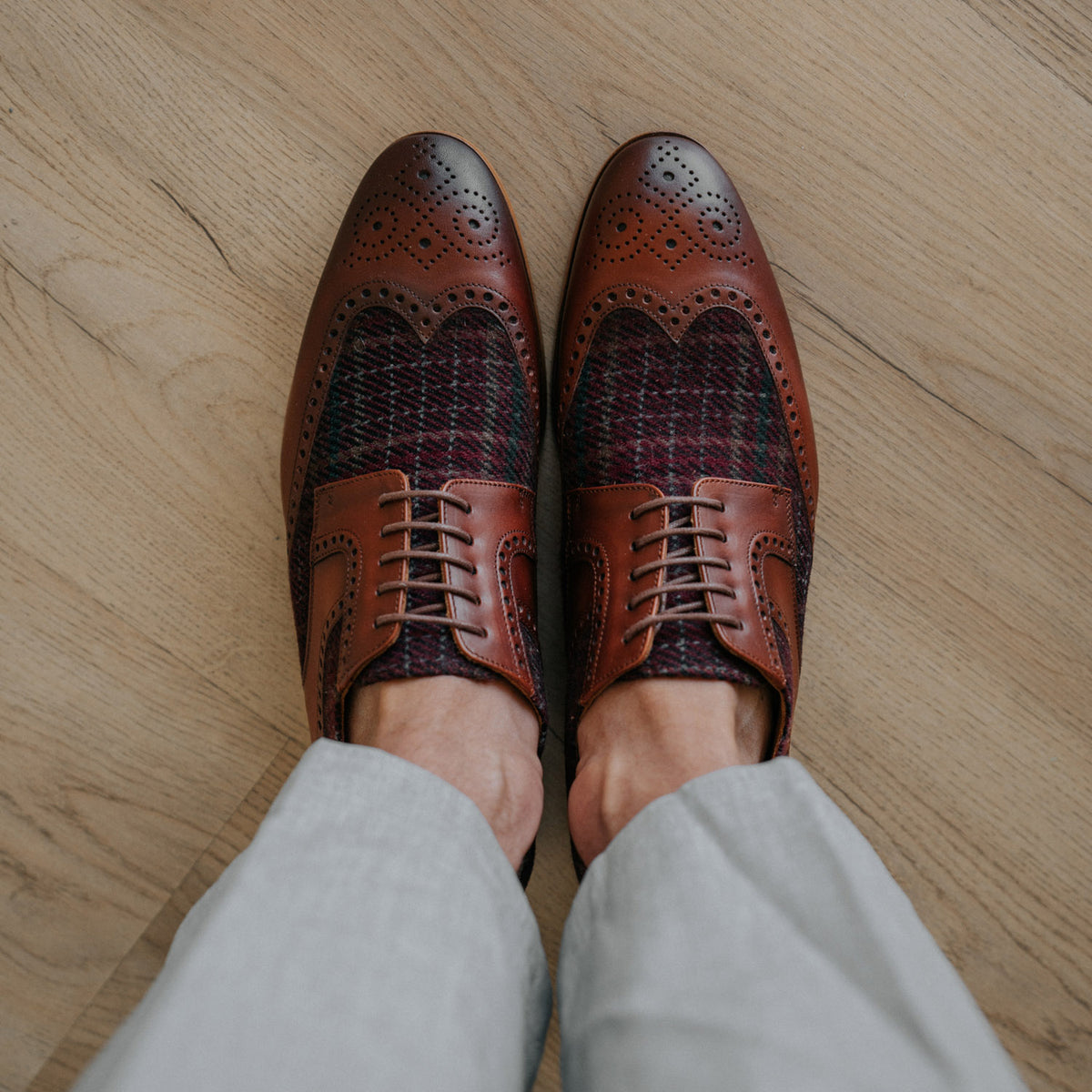 Brown leather and tweed oxford shoes with brogue detailing on a wooden floor, worn by a person in light gray pants.