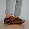 A person wearing light gray trousers and brown leather and fabric brogue shoes stands on a wooden floor.