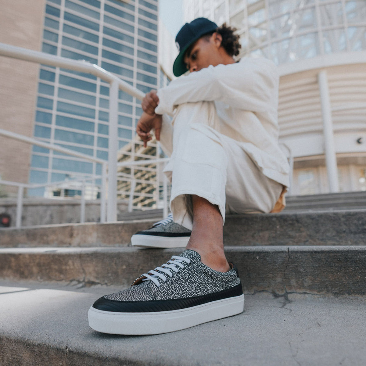 A person in white clothing and a dark cap sits on outdoor steps, wearing patterned sneakers with white laces and thick white soles. A modern building facade is visible in the background.