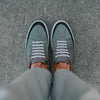 Person wearing grey shoes with white laces, standing on a textured grey surface. The view is from above, showing their lower legs and feet.