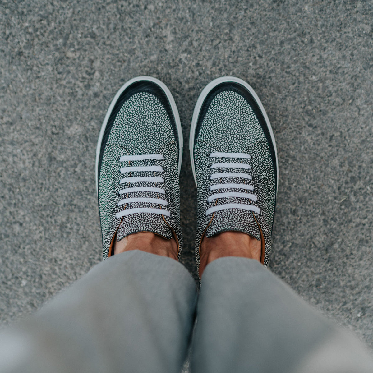 Person wearing grey shoes with white laces, standing on a textured grey surface. The view is from above, showing their lower legs and feet.