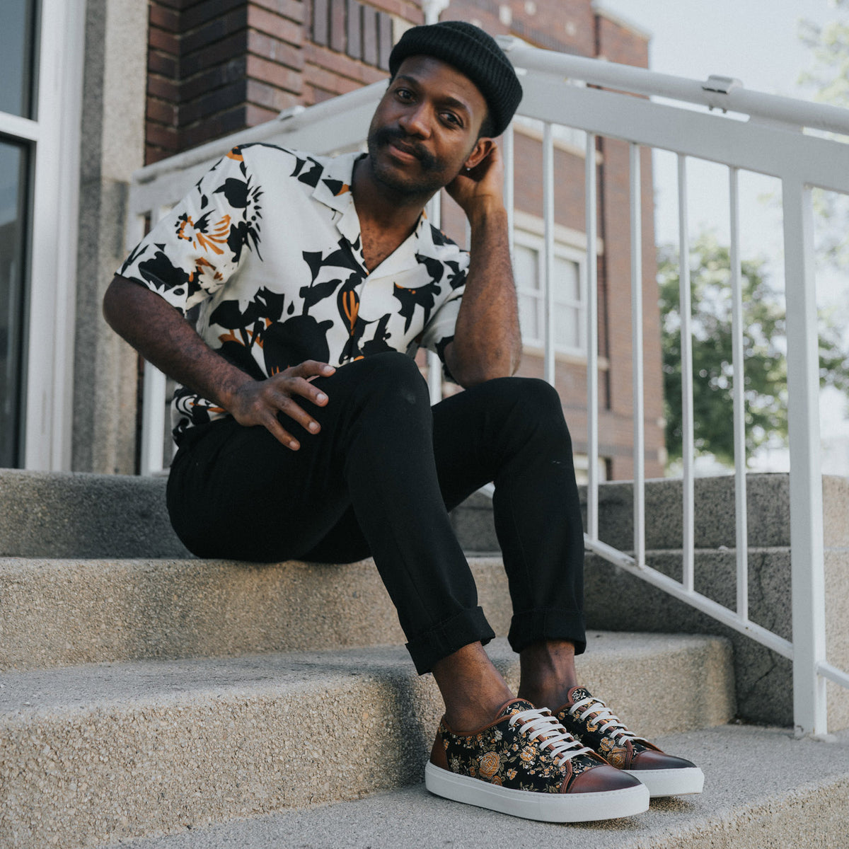 A person sits on outdoor concrete steps, wearing a patterned shirt, black pants, and floral-patterned sneakers while resting their hand on the side of their head.