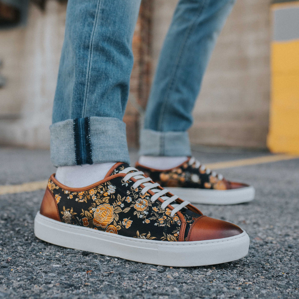 Person wearing floral-patterned sneakers with rolled-up blue jeans, standing on pavement.