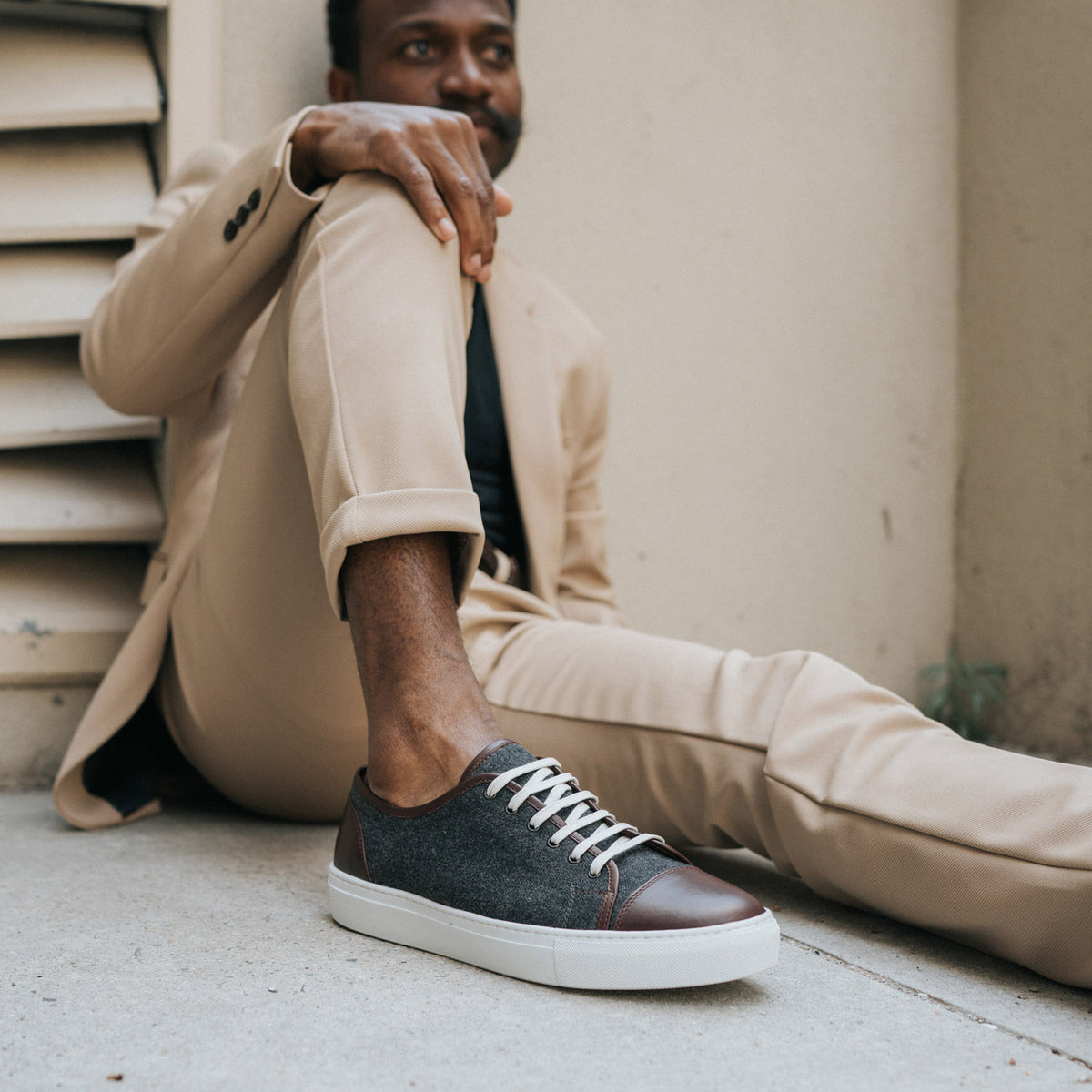 Person wearing a beige suit and dark gray sneakers with white laces, sitting on a concrete floor against a wall.