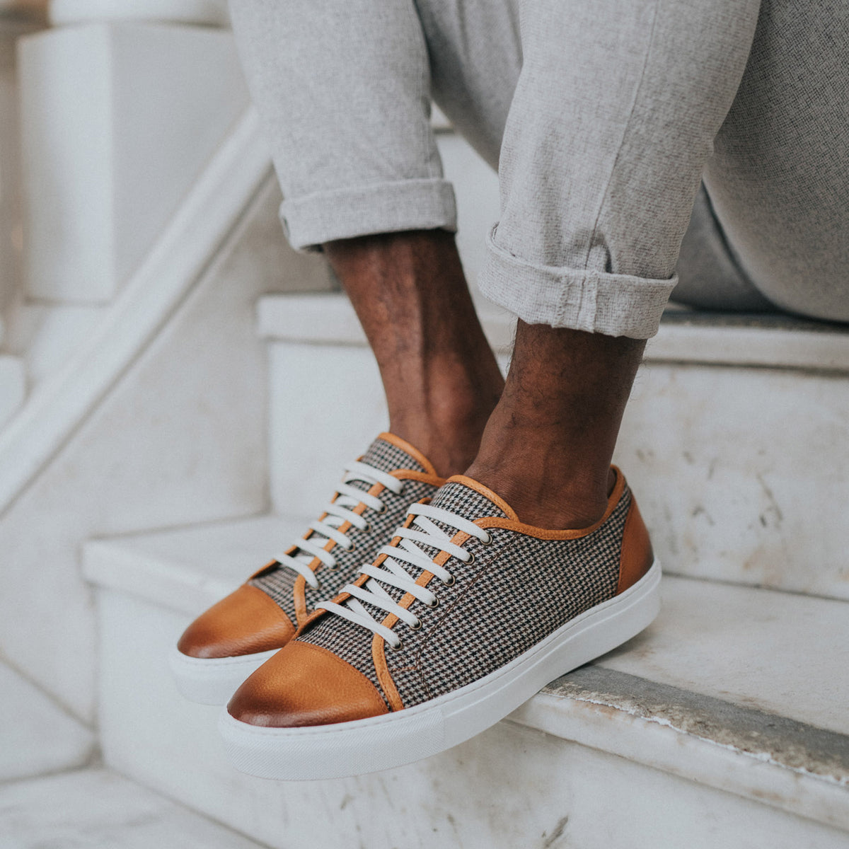 Close-up of a person sitting on marble stairs wearing grey pants and stylish sneakers with a mix of brown leather and grey fabric and white laces.