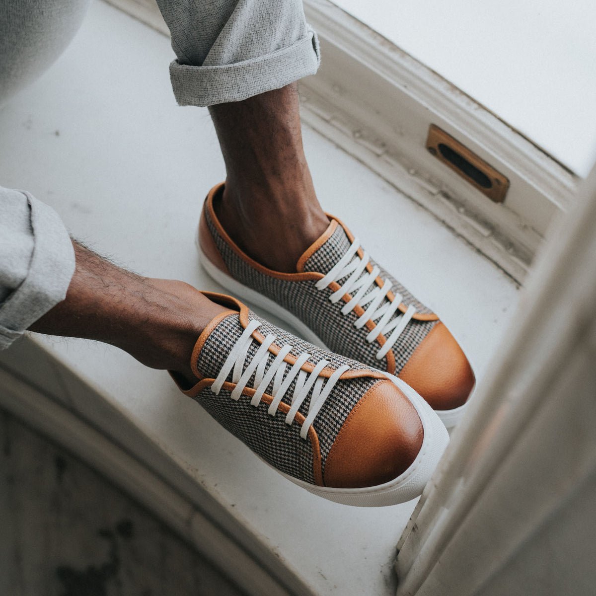 A person wearing grey and orange sneakers with white laces, sitting on a windowsill. The person is dressed in cuffed grey pants.