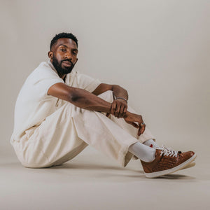 A man sits on the floor against a neutral background, wearing a white shirt, beige pants, white socks, and brown sneakers.