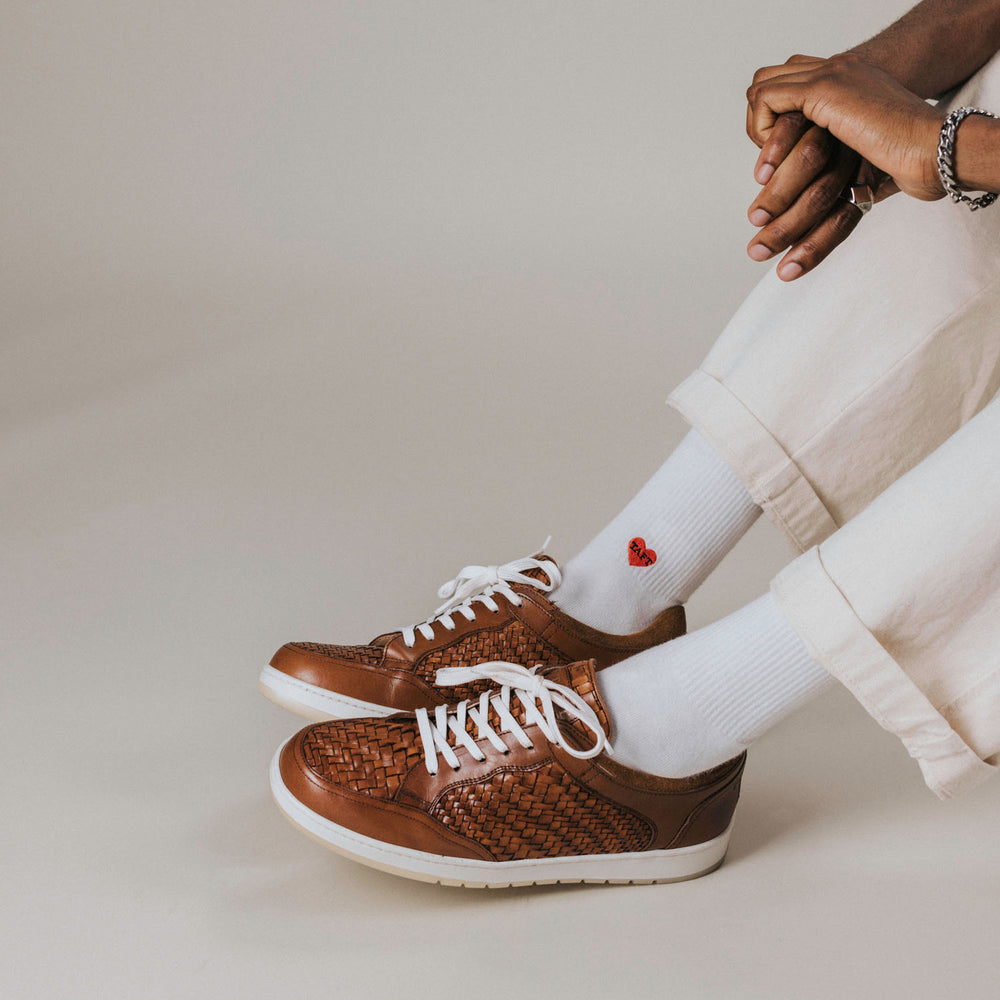 A person wearing white pants and white socks with small red heart embroidery, sitting with legs crossed. They are wearing brown woven leather sneakers with white laces.