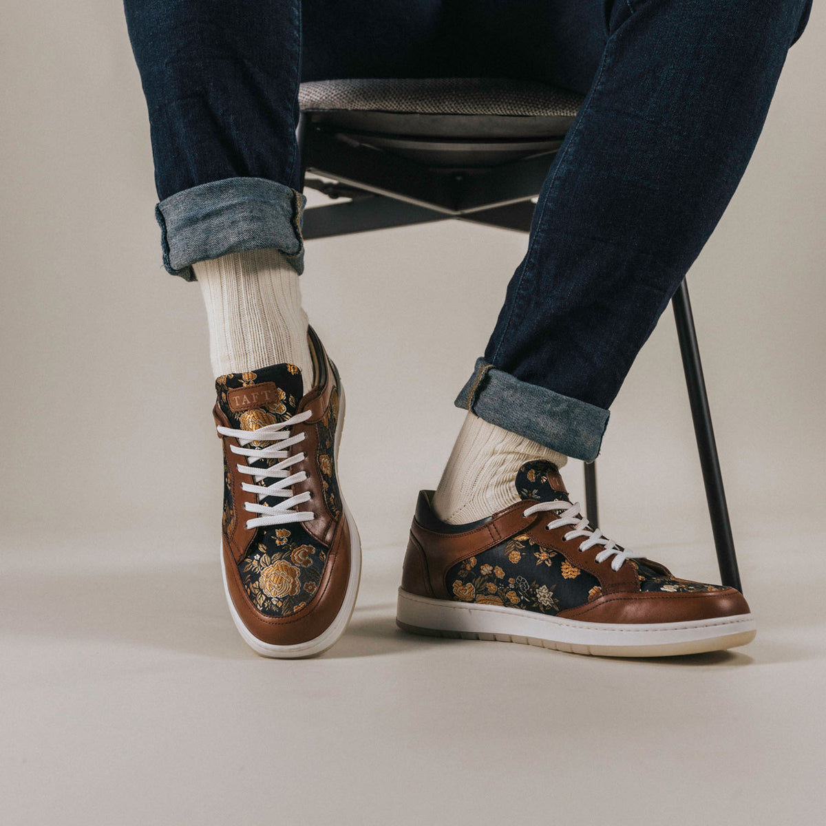 Person wearing floral-patterned sneakers and white socks while sitting on a chair, with jeans rolled up at the ankles.