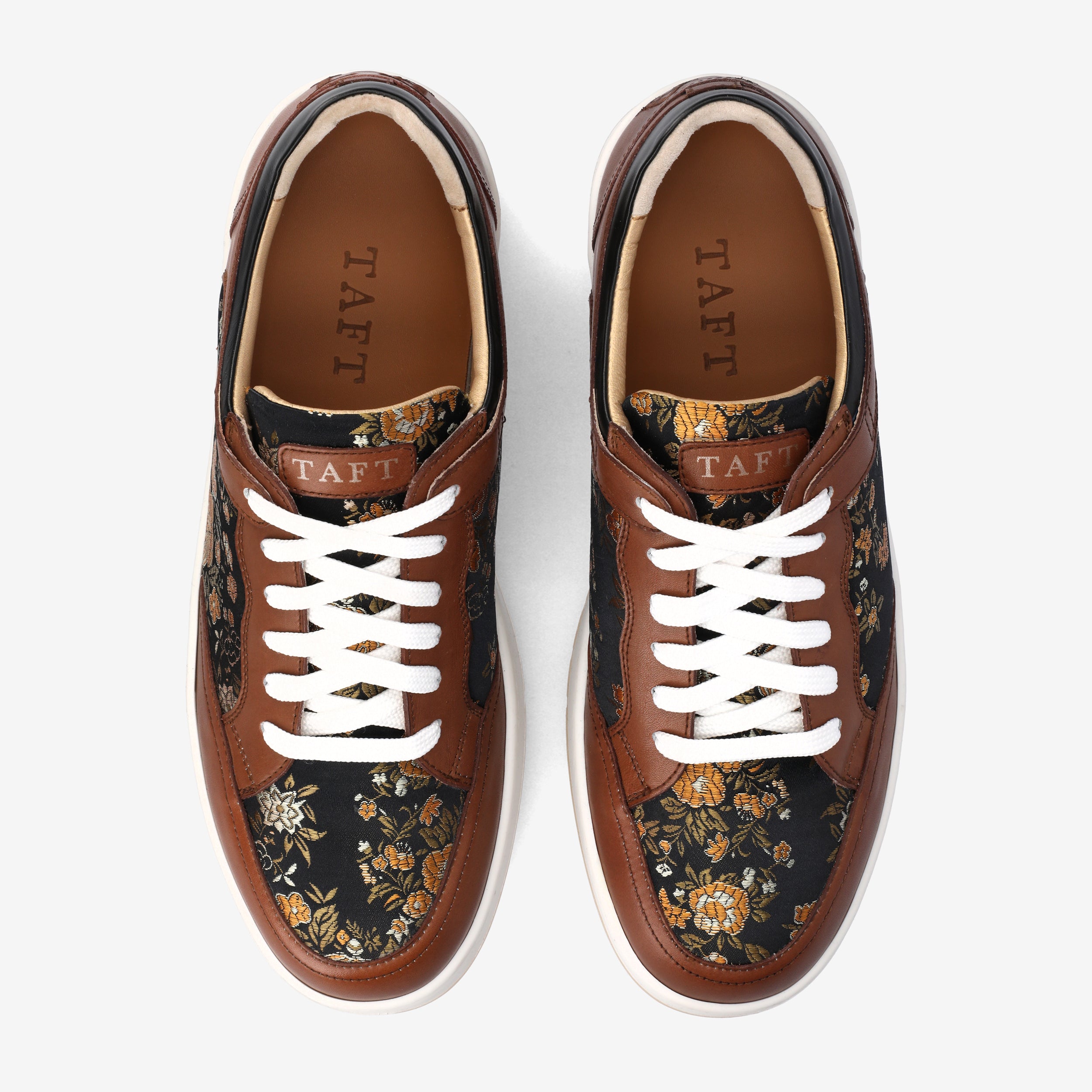 A pair of low-top sneakers featuring brown leather overlays, floral-patterned fabric, and white laces is displayed from a top-down view. The brand name TAFT is visible on the tongue and inside the shoe.