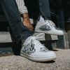 A person wearing white and floral-patterned sneakers, white socks, and distressed black jeans sits on concrete steps.