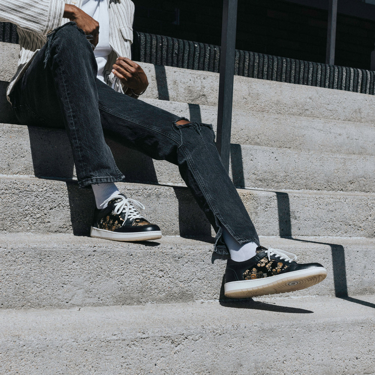 Person wearing black floral-patterned sneakers, white socks, ripped black jeans, and a white jacket sits on concrete steps.