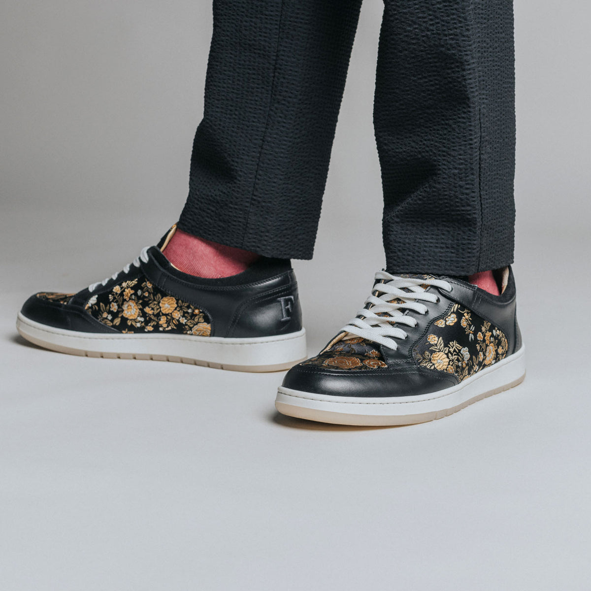 A person wearing black pants and black leather sneakers with gold and bronze floral embroidery and white laces.