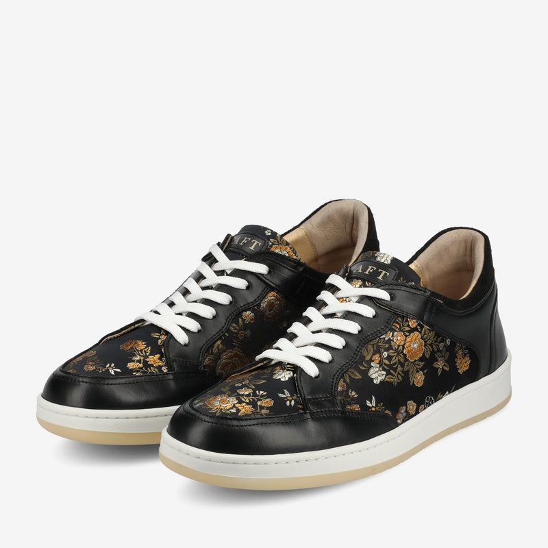 A pair of black sneakers with white laces, featuring a floral pattern with yellow and brown flowers on the sides and front, and white soles.