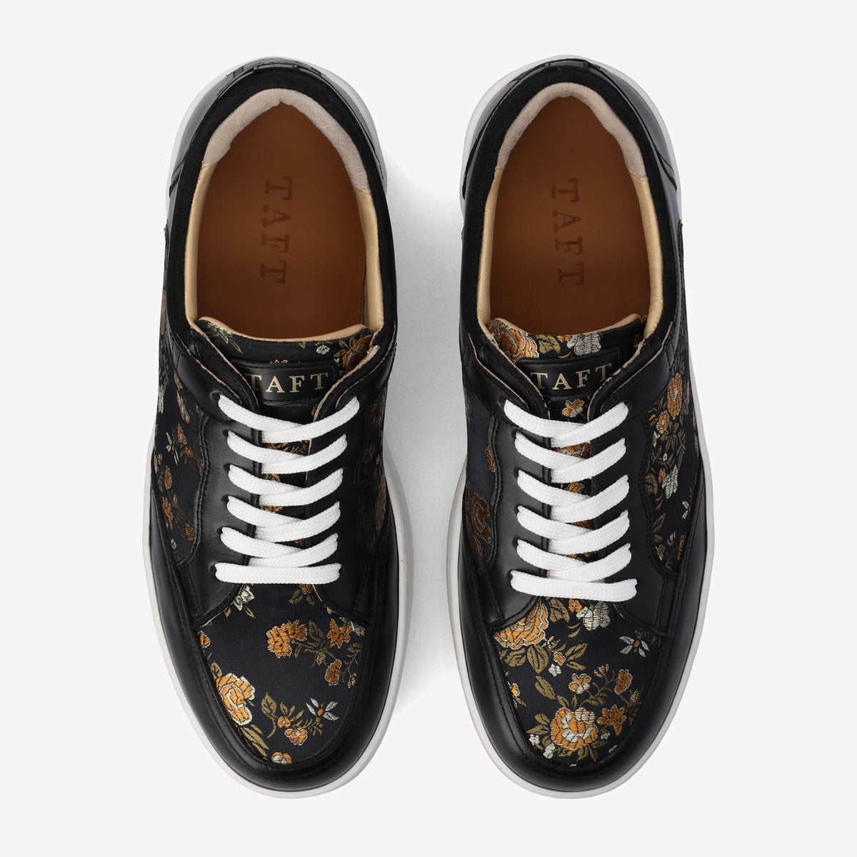 A pair of black sneakers with floral patterns and white laces, viewed from above. The inner soles are brown with the brand name TAFT visible.